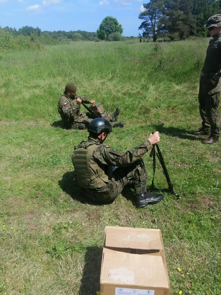 Kadet w hełmie i kamizelce taktycznej podczas zajęć w terenie, w tle instruktorzy