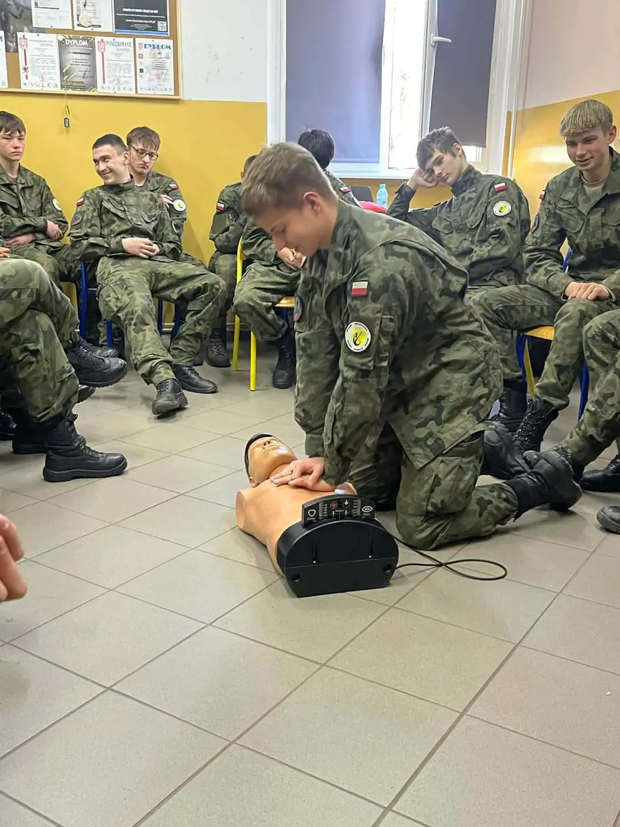Kadet uciska klatkę piersiową fantoma, obok grupa kadetów na krzesłach