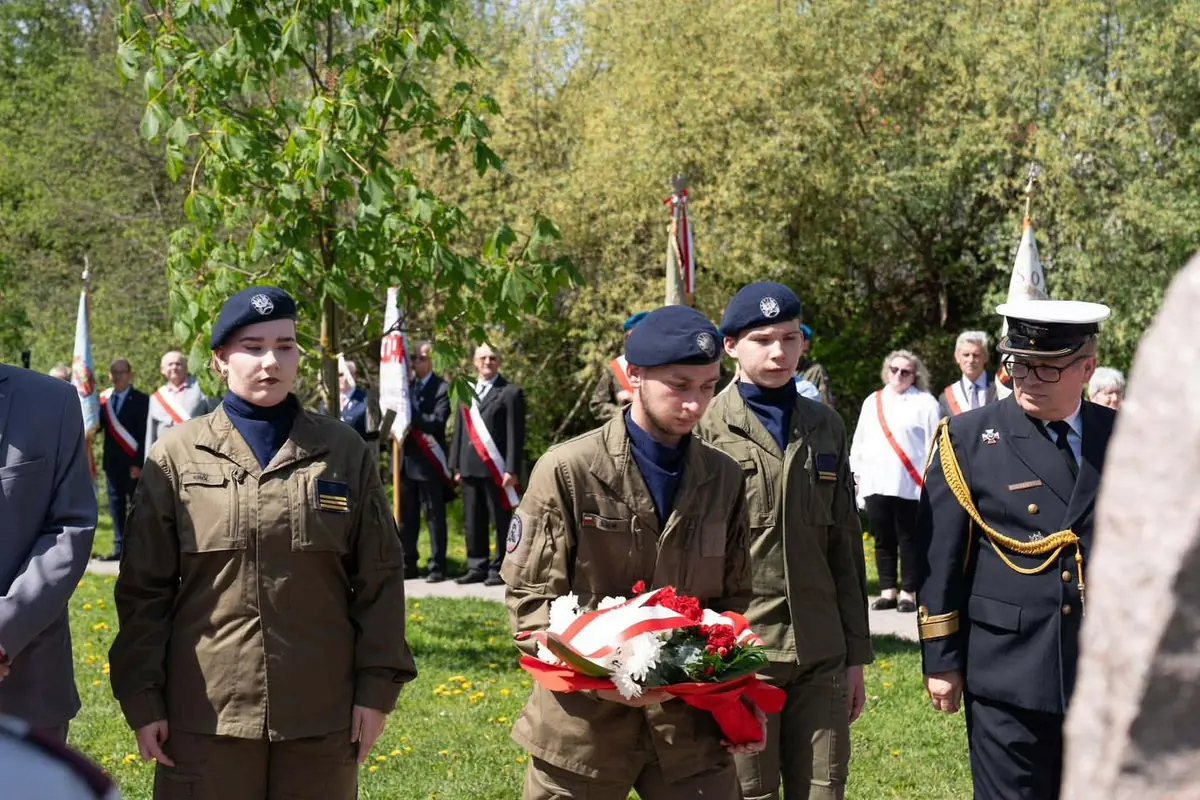 Kadet niesie wiązankę kwiatów przewiązaną biało-czerwoną wstęgą, obok kadeci w beretach