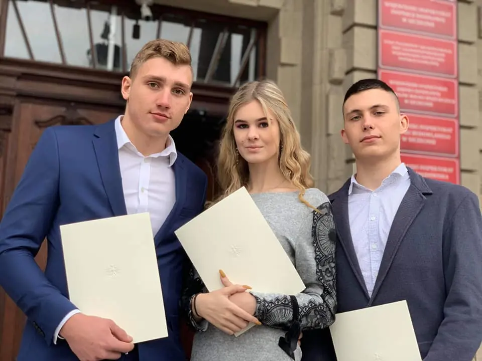 Troje uczniów w strojach galowych z teczkami dyplomów przed budynkiem urzędu